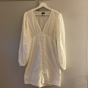 Vit broderad klänning Vero Moda - Adoore liknande klänning från Vero Moda med broderade blommor, v-ringning och klädda knappar framtill. Långa puffärmar med resår vid handlederna och lätt A-linjeformad passform. Perfekt för sommarens alla tillfällen!