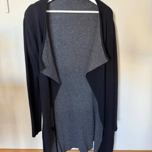 Svart och grå cardigan jacka - Säljer en svart och grå långärmad cardigan jacka med öppen front och mjukt, stickat material. Jackan har en snygg tvåfärgad design med mörk utsida och ljusare grå insida. Perfekt för lager-på-lager och enkel att matcha till olika outfits.