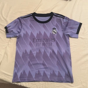 Real Madrid bortatröja 2022/23 - Säljer en lila Real Madrid bortatröja från Adidas med svart krage och svarta detaljer på ärmarna. Tröjan har ett grafiskt mönster och klubbmärket på bröstet samt sponsortryck. Perfekt för dig som älskar fotboll och Real Madrid.