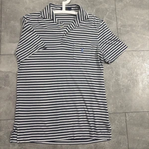 Randig pikétröja från Polo Ralph Lauren - Säljer en klassisk vit och marinblå randig pikétröja från Polo Ralph Lauren. Tröjan har korta ärmar, krage och knappslå samt en liten broderad logga på bröstfickan. Perfekt för en stilren look.