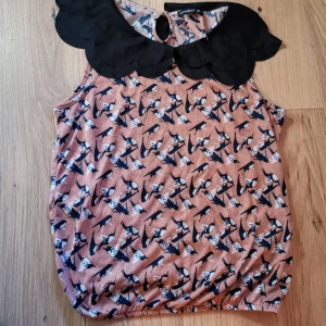 Mönstrad ärmlös blus med svart krage - Säljer en söt ärmlös blus med fågelmönster i svart och vitt på en rosa/beige botten. Blusen har en stor dekorativ svart krage med vågig kant och knapp i nacken. Perfekt för dig som vill sticka ut lite extra!Den passar storlek 42. Den har ytludd som den fick snabbt 