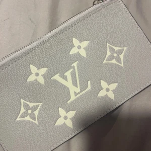 Grå kuvertväska - Säljer en stilren grå kuvertväska med klassiskt monogramtryck i vitt. Väskan har dragkedja upptill och är tillverkad i ett strukturerat material som ger en lyxig känsla. Perfekt att bära till alla tillfällen.