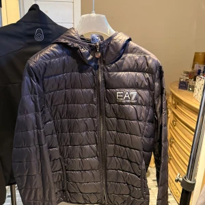Svart lättviktsjacka med huva från EA7 och en Svart Sail racing jacka med hög krage  - Säljer en svart pufferjacka från EA7 Emporio Armani. Jackan har huva, dragkedja framtill och EA7-logga på bröstet. Perfekt för kyliga dagar och enkel att bära med sig tack vare lättviktsmaterialet. Och även en riktig fin sail-racing jacka svart med hög krage 