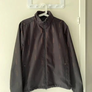 Svart vindjacka med diskret mönster från J Lindberg - Säljer en svart vindjacka med diskret mönster och dragkedja framtill. Jackan har hög krage och två fickor med dragkedja. Perfekt för blåsiga dagar och enkel att matcha med det mesta.