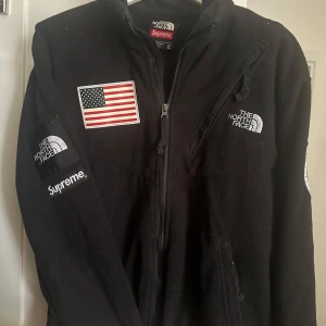 Svart fleecejacka från The North Face x Supreme - Svart fleecejacka med broderade loggor från The North Face och Supreme. Amerikansk flagga på bröstet och ryggen. Jackan har dragkedja framtill och flera fickor med dragkedja. Fick denna av ASAP Rocky när han var i stockholm.