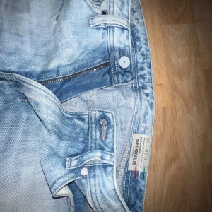 Ljusblå jeansbyxor från Diesel - Säljer ett par ljusblå jeansbyxor från Diesel med klassisk femficksdesign och snygga slitningar. Byxorna har normal passform och dragkedja i gylfen. Perfekta för dig som gillar en avslappnad stil.