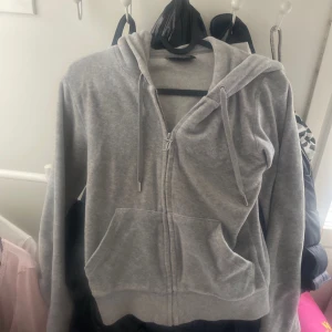 Grå hoodie från Juicy Couture - Mjuk och klassisk grå hoodie från Juicy Couture med dragkedja framtill och två fickor. Tröjan har huva med snören och ribbade muddar vid ärmslut och nederkant. Perfekt för en avslappnad stil.