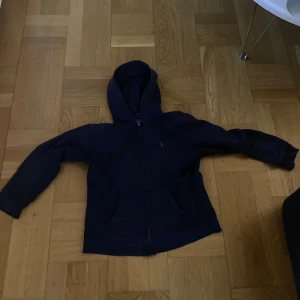 Mörkblå hoodie från Polo Ralph Lauren - Säljer en mörkblå hoodie från Polo Ralph Lauren med dragkedja och huva. Tröjan har två fickor framtill och den klassiska röda loggan broderad på bröstet. Perfekt för en avslappnad stil.