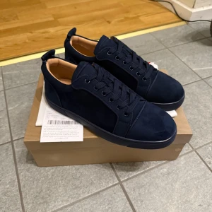 Mörkblå loubs storlek 43 - Säljer ett par mörkblå sneakers från Christian Louboutin i mocka med klassisk röd sula. Skorna har rund tå, platt sula och snörning. Perfekta för dig som vill ha en stilren och lyxig look.
