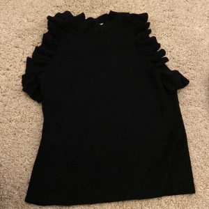 Svart ribbad topp med volang från H&M - Säljer en svart ribbad topp från H&M med volangdetaljer längs axlarna. Toppen är ärmlös och har en rund halsringning. Perfekt för att ge outfiten lite extra edge!