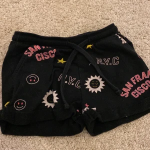 Svarta shorts med tryck från DREAMS - Svarta shorts med färgglada tryck som 'SAN FRANCISCO', 'N.Y.C', glada smileys och solmotiv. De har snörning i midjan och är perfekta för en avslappnad stil. Mjukt material och bekväm passform.