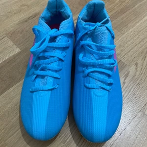 Blå adidas fotbollsskor med snörning - Säljer ett par blå adidas fotbollsskor med snörning och vita dobbar. Skorna har rosa adidas-logga på sidan och insidan är fodrad i blått och rosa. Perfekta för spel på gräsplan.