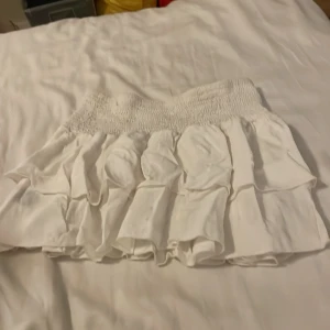 Vit volangkjol från SHEIN - Supersöt vit kortkjol från SHEIN med volanger och smock i midjan. Kjolen är luftig och har en feminin look, perfekt för varma dagar. Modellen är Petite XS och passar dig som gillar en lätt och flowy stil.