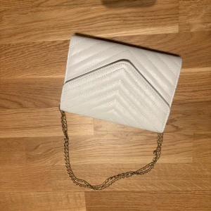 Vit quiltad axelväska med guldkedja - Säljer en stilren vit axelväska med quiltat mönster och guldfärgad kedja. Väskan har ett innerfack med dragkedja och stängs med lock. Perfekt för att lyfta din outfit!