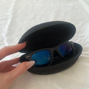 Svarta solglasögon från Polaroid - Snygga svarta solglasögon från Polaroid med blåtonade spegelglas. Klassisk wayfarer-form och medföljande svart hårt fodral. Perfekta för soliga dagar och ger en cool look.