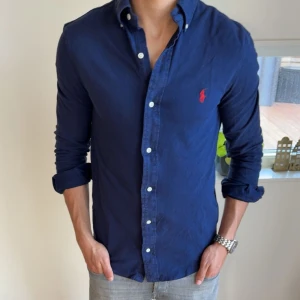 Ralph Lauren skjorta - Säljer en klassisk mörkblå skjorta från Ralph Lauren med knappar framtill och röd broderad logga på bröstet. Skjortan har lång ärm och button-down krage, perfekt till jeans eller chinos.