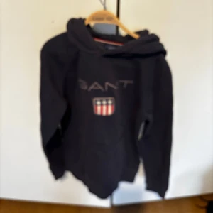 Mörkblå hoodie från Gant - Säljer en mörkblå hoodie från Gant med klassisk logga och sköld på bröstet. Tröjan har huva och ribbade muddar vid ärmslut och nederkant. Perfekt för dig som gillar stilrena och sportiga plagg.