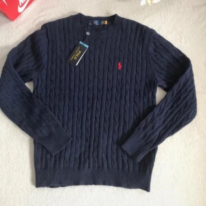 Mörkblå stickad tröja från Polo Ralph Lauren - Säljer en mörkblå stickad tröja från Polo Ralph Lauren med klassiskt kabelstickat mönster och rund halsringning. Tröjan har lång ärm och den ikoniska röda loggan broderad på bröstet. Perfekt för en stilren och tidlös look.