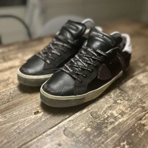 Svarta sneakers från Philippe Model - Säljer ett par svarta sneakers från Philippe Model med vita detaljer och svart snörning. Skorna har en platt sula och är tillverkade i skinn med en klassisk design. Perfekta för dig som gillar stilrena och bekväma skor. Pris kan sänkas vid snabb affär