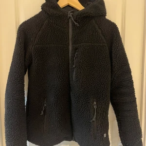 Svart fleecejacka med huva från Brandit - Mysig svart fleecejacka från Brandit med huva och dragkedja framtill. Jackan har tre fickor med dragkedja och är perfekt för kyliga dagar.  I storlek Xs s kan också passa 