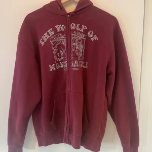 Vinröd hoodie med dragkedja från One of One - Vinröd hoodie med dragkedja från One of One. Tröjan har huva, fickor framtill och ett tryck med text och motiv på bröstet. Perfekt för en avslappnad stil.