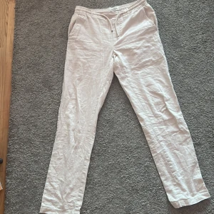 Beiga linnebyxor från Dressmann med snörning - Säljer ett par beiga linnebyxor från Dressmann i storlek S. Byxorna har elastisk midja med snörning och sidfickor, samt bakfickor med knapp. Perfekta för varma dagar och en avslappnad stil.