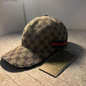Beige keps från Gucci  - Säljer en beige keps från Gucci med klassiskt GG-monogram och röd-blå rand på sidan. Justerbar rem baktill i skinn. Snygg och tidlös design som passar till många stilar. Storlek ”M”