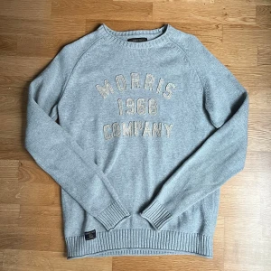Stickad tröja från Morris - Säljer en ljusgrå stickad tröja från Morris Gents med broderad text 'Morris 1968 Company' på bröstet. Tröjan har långa ärmar och ribbade muddar vid ärmslut och nederkant. Perfekt för en stilren och avslappnad look till sommaren.