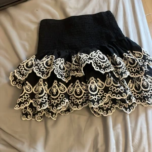 Svart volangkjol med broderade detaljer från Lindex - Supersöt svart kjol från Lindex med bred smock i midjan och två lager volanger. Kjolen har vita broderade mönster längs volangerna som ger en unik och lekfull look. Perfekt för dig som vill sticka ut lite extra. Den har ett extra lager innanför! storlek 134-140!Den är stretchig