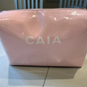 Ljusrosa necessär från CAIA - Säljer en ljusrosa necessär från CAIA med dragkedja upptill. Väskan har en glansig yta och är perfekt för att förvara smink eller andra småsaker. Enkel och stilren design med CAIA-logga på framsidan. Använd men i fint skick. Nytvättad
