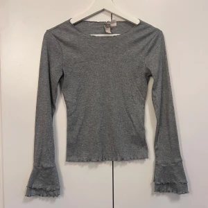 Grå ribbad långärmad topp med volangärm  - Säljer en grå ribbad långärmad topp från  H&M. Toppen har volangdetaljer vid ärmslut och nederkant samt en tight passform. Perfekt att styla med jeans eller kjol för en trendig look.