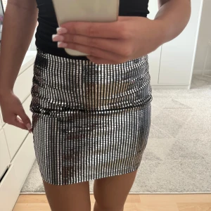 Glittrig silverkjol från New Yorker - Säljer en glittrig, tight minikjol från New Yorker i silver. Kjolen har ett rutigt mönster av små spegel-liknande paljetter. Ny med prislapp i storlek 34