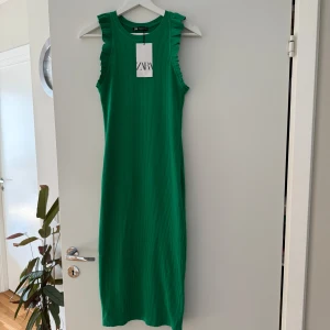 Ny grön ribbad klänning från Zara. S - Säljer en grön ribbad klänning från Zara i storlek S. Klänningen är ärmlös med volangdetaljer vid axlarna och har en rak, kroppsnära passform som slutar under knäet(112 cm lång). Perfekt för dig som gillar färg och stilrena detaljer. Djurfritt och rökfritt hem.