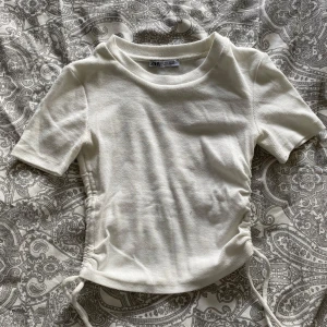 Vit ribbad topp med knyt från Zara - Säljer en vit ribbad topp från Zara i storlek S. Toppen har korta ärmar och knyt i sidorna som ger en snygg detalj och möjlighet att justera passformen. Perfekt till vår och sommar!