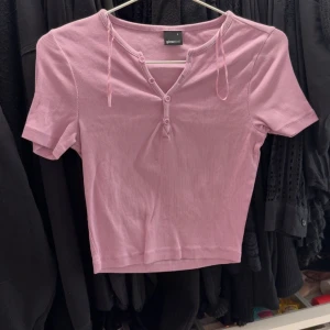 Lila ribbad topp  - Säljer en lila ribbad topp från gina med korta ärmar och knappar framtill. Toppen har v-ringning och dekorativa knytband vid halsen. Perfekt för en trendig och avslappnad stil.