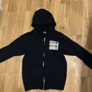  hoodie med dragkedja  - Svart hoodie med dragkedja  Klassisk rutig detalj på bröstet i beige, svart, vit och röd. Huva och fickor framtill. Perfekt för en stilren och avslappnad look.