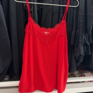 Röd linnetopp med spets - Säljer en röd linnetopp med tunna axelband och vacker spetsdetalj vid urringningen. Perfekt för att addera färg till din outfit. Toppens material känns mjukt och lätt.