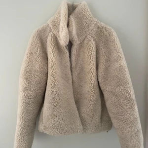 Beige teddyjacka med hög krage - Mysig beige teddyjacka med hög krage och rak passform. Jackan är tillverkad i ett fluffigt teddy-material som håller dig varm under kyliga dagar. Perfekt för dig som vill ha en trendig och bekväm look.