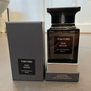 Tom Ford Oud Wood Eau de Parfum 100 ml - Säljer en elegant och lyxig parfymflaska av Tom Ford Oud Wood, Eau de Parfum, 100 ml. Flaskan är mörk och stilren med en fyrkantig form och ett svart lock. Perfekt för den som älskar exklusiva dofter med en touch av oud.