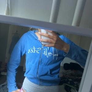 Blå hoodie från Hollister - Säljer en blå hoodie från Hollister med vit broderad logga på bröstet. Tröjan har dragkedja framtill, huva med snören och ribbade muddar. Perfekt för en avslappnad stil.