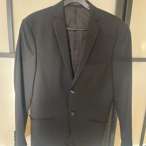 Svart kostym + byxor från Tiger of Sweden - Säljer en klassisk svart kostym + kostymbyxor från Tiger of Sweden. Kavajen har två knappar, smal passform och diskreta fickor. Byxorna är raka och matchar kavajen perfekt. Kavajen och byxorna är köpta tillsammans från tiger of Sweden för ungefär 4500kr. Kavajen och byxorna har endast använts under en avslutning då jag växt ur dem. Strlk S. Hör av dig vid fler frågor eller bilder!