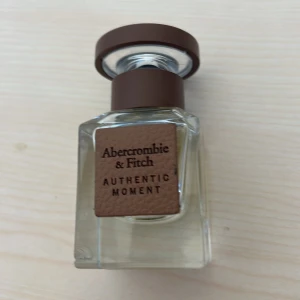Abercrombie & Fitch Authentic Moment parfym - Säljer en stilren parfym från Abercrombie & Fitch, Authentic Moment. Flaskan är fyrkantig i genomskinligt glas med ett brunt lock och en brun etikett framtill. Perfekt för dig som gillar eleganta och moderna dofter.