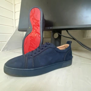 Christian Louboutin skor - Säljer ett par mörkblå sneakers från Christian Louboutin med ikonisk röd sula och diskret logga på insidan. Skorna har snörning och rund tå, samt en mjuk innersula i beige läder. Perfekta för dig som vill sticka ut med stil.(INTE ÄKTA)