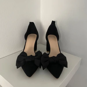 Svarta klackskor med rosett - Säljer ett par eleganta svarta klackskor med spetsig tå och stor dekorativ rosett framtill. Skorna har en stabil klack och är perfekta för att lyfta din outfit💗