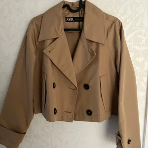 Beige croppad trenchcoat från Zara - Säljer en beige croppad trenchcoat från Zara med dubbelknäppning och bred krage. Jackan har långa ärmar och två fickor framtill. 