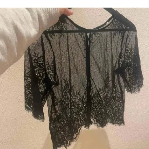 Svart spetsblus med kort ärm - Säljer en transparent svart spetsblus med korta ärmar och fina spetsdetaljer. Blusen har knäppning framtill och vacker mönstrad spets längs kanterna. Perfekt för att lyfta en outfit med en elegant touch.