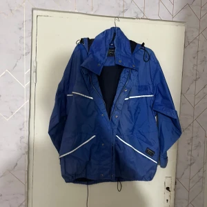 Blå vindjacka från Norman Wells - Säljer en blå vindjacka från Norman Wells Sportswear med huva och vita reflexdetaljer. Jackan har dragkedja framtill, flera fickor och justerbar nederkant. Perfekt för blåsiga dagar och utomhusaktiviteter.