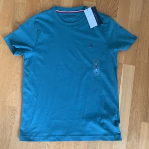Turkos t-shirt från Tommy Hilfiger - Säljer en turkos t-shirt från Tommy Hilfiger med liten broderad logga på bröstet. Klassisk rund halsringning och korta ärmar. Perfekt basic-plagg med snygg passform och diskret branding.