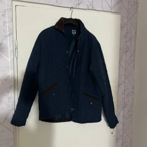 Mörkblå quiltad jacka från EP Collection - Säljer en mörkblå quiltad jacka från EP Collection med bruna detaljer vid krage och fickor. Jackan har lång ärm med förstärkta armbågar och knäppning framtill. Perfekt för kyligare dagar.