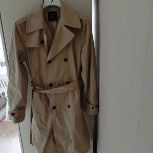 Beige trenchcoat från Boomerang Herr Storlek M  - Klassisk beige trenchcoat från Boomerang med dubbelknäppning, bred krage och bälte i midjan. Jackan har långa ärmar och knappar framtill, perfekt för en stilren look under vår och höst.Galge ingår ej.Nypris 3600 kr.